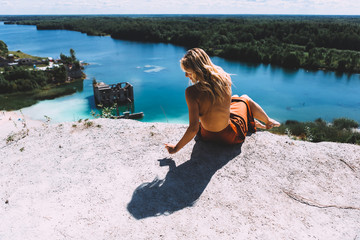 At a picturesque vantage point of Rummu quarry, a blond woman is standing in her bare feet, wearing a thigh high, auburn coloured dress, With her back turned away from the camera. Clear Water.