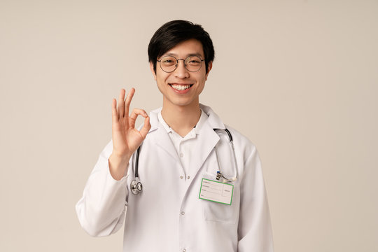 Image of asian young male doctor in uniform smiling and gesturing ok sign