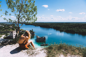 At a picturesque vantage point of Rummu quarry, a blond woman is sitting, wearing a thigh high, auburn coloured dress, With her back turned away from the camera. Clear Water.