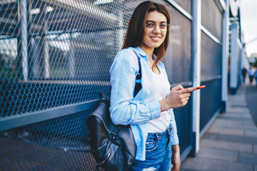 Half length portrait of cheerful caucasian woman in spectacles posing on urban setting while walking in city with smartphone,smiling hipster girl in stylish apparel chatting via mobile phone on street