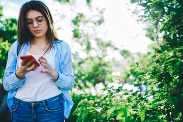 Fototapeta premium Pensive caucasian millennial woman standing on green trees background in park holding mobile phone, young brunette female blogger checking mail spending free time on sunny day outdoors