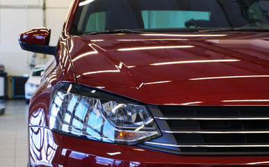 Front view of a modern luxury car in bright red. Pictured: headlight, part of the grille and hood. New car sales, service and spare parts. Concept.
