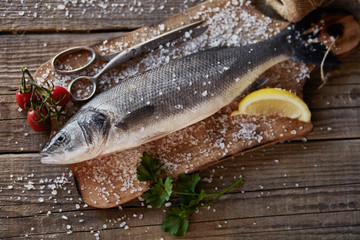 Sea bass fish on the dark wooden background with salt, tomatoes cherry and lemon