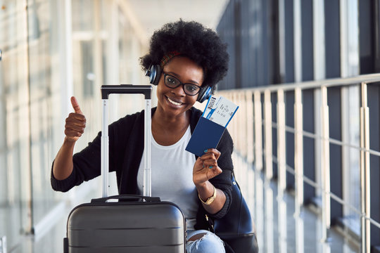 Young african american female passanger in casual clothes and headphones is in airport with baggage