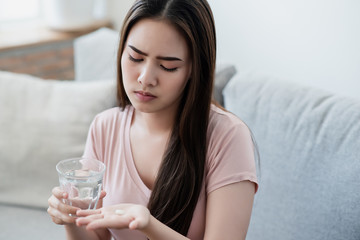 Sick asian woman eating pills with a glass of water in hand with tired face. Healthy, medicine taking, chemical, illness, sickness, health care, pharmacy, virus, covid 19 prevent concept.