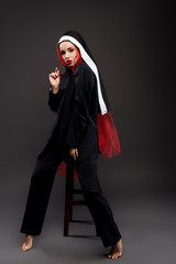 barefoot fashionable sexy nun posing in black suit and red scarf with sunglasses, on grey