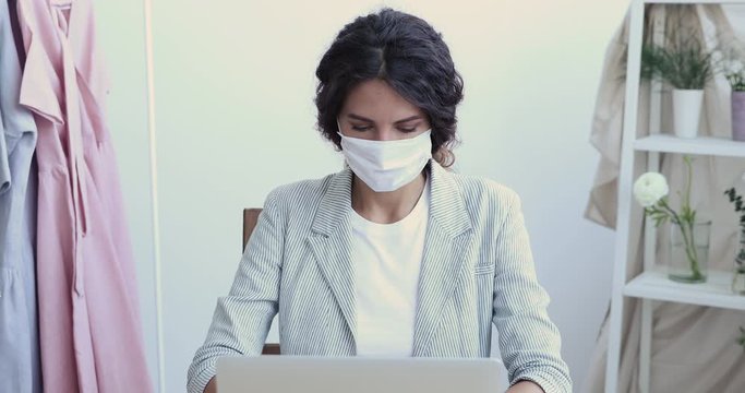 Female fashion designer working distantly from home office using laptop. Young businesswoman wearing face mask typing on computer doing remote job during coronavirus pandemic quarantine concept.