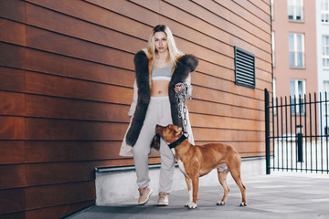 Long haired blonde, wearing a sport suit, a white jacket with fur. Gracefully holding a pitbull by...