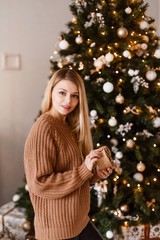 Blonde in a brown knitted sweater holds a gift in her hands next to a decorated Christmas tree, holding present. Christmas photo