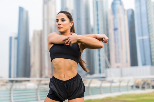 Young fitness woman make warming hands exercises before run against skyscrapers background