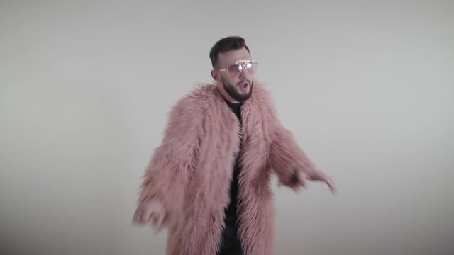 Portrait of cheerful homosexual Caucasian man dancing on white background. Handsome brunette guy in pink fur coat and sunglasses having fun. Lifestyle, sexual orientation, joy, happiness.