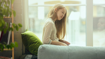 Fototapeta premium Little girl sitting near by window with tablet, remote studying, being at home. Doing homework, online serfing, chatting, conference with teacher. Staying home, indoors lifestyle, education concept.
