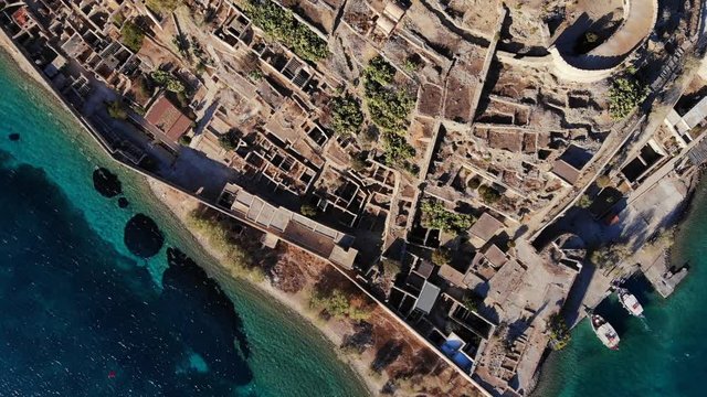 Top view of Spinalonga fortress, old buildings and defence structures ruins on rocky island. Tourist boats moored at pier, land area filled with remains of ancient constructions