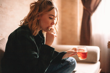 Young thoughtful upset woman drinking tea and eating chocolate while sitting on sofa in living room at home