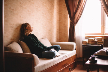 Young upset woman holding book looking through window while sitting on sofa in living room at home - Powered by Adobe