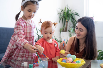 Happy young beautiful mother spending time with her joyful daughters. Cheerful family mom and children painting easter eggs with colors. Little pretty girl putting bunny ears. Preparation for Easter.