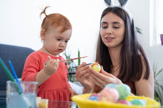Attractive young woman with little cute baby girl are preparing for Easter celebration. Mom and daughter wearing bunny ears are spending time together before Easter while painting eggs.