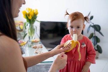 Cute red hair baby girl in red dress eating banana. Pretty beautiful funny kid with fruits. Yellow flowers background. White light.