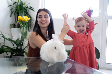 Cheerful smiley mother and her daughters playing with Easter white rabbit and laughing. Happy family preparing for Easter. Easter family concept. White window background. Yellow flowers.