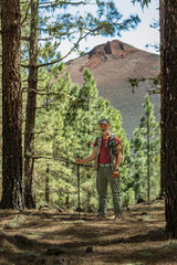 Traveler in a cap with backpack, hiking sticks has stopped to make a shot. Pine tree forest near the Volcano Arenas Negras. Blue sky and the top of volcano in the background. Tenerife, Canary Islands