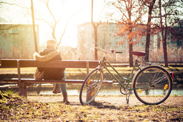 Fototapeta na wymiar Enjoying the sun in spring: Young girl is sitting on park bench, bicycle back view
