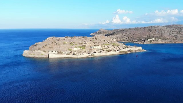 Spinalonga and Spinalonga peninsula, aerial shot, camera fly back, away from famous island. Calm sea waters surround former fortifications on small isle at Lasithi, North-East Crete Island