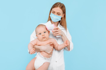 a female pediatrician in a medical mask with a digital infrared thermometer, measures the body temperature of the baby