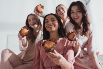 Selective focus of multicultural women showing muffins, smiling and looking at camera at bachelorette party