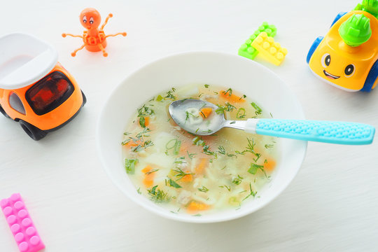 .White plate. with soup and a spoon with a blue plastic handle. Chicken stock soup with curly noodles, carrots and herbs. Different bright toys..