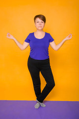 Woman at the age of doing yoga standing on a yellow background.