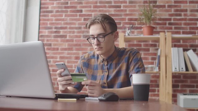 A young hipster enters Bank card details into his phone while sitting at his Desk with a laptop, computer mouse, stickers and coffee. Student makes online purchases