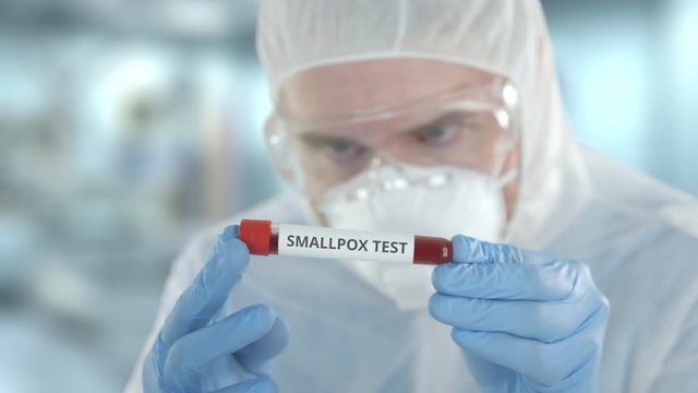 Defocused laboratory assistant wearing protection suit examines vial with smallpox test