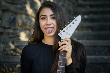 Portrait of a young woman looking forward and sticking out her tongue, dressed in a sweater and black trousers and holding a white electric guitar