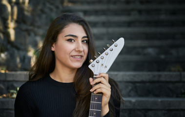 Portrait of a young woman facing the front, dressed in a sweater and black trousers and holding a white electric guitar
