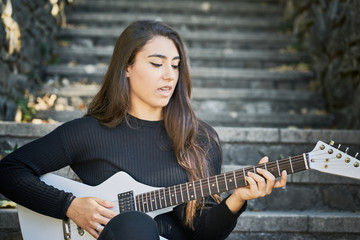 young woman dressed in a sweater and black trousers and playing a white electric guitar sitting on a stone staircase