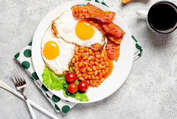 Fried eggs, crispy bacon, beans and tomatoes in a white plate on a gray concrete background, top view, copy space. Breakfast with eggs, bacon and coffee, free space for text.