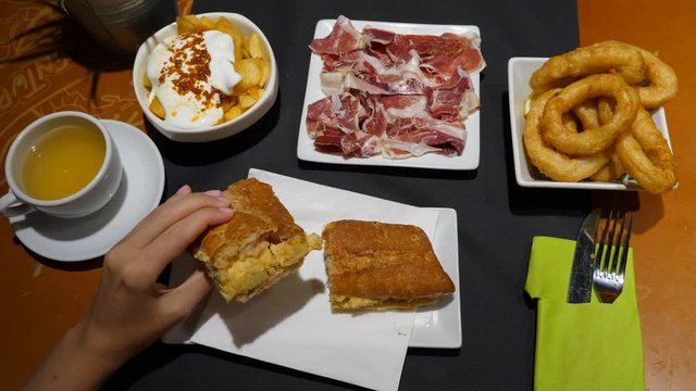 Typical food at Spanish restaurant, top view of table with potato, jamon, fried calamares and potato omelette sandwich. Woman hand take up piece of bread