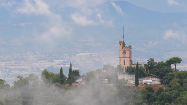 White clouds tear to patches and fly low over mountain with tower building on top, time lapse shot. Telephoto perspective of Turo del Puig. Standalone villa surround with forested area of Sant Cugat