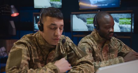 Multiethnic military men in uniforms and headsets talking and videochatting while working as dispatchers. Soldiers sitting at computers and speaking in controlling room.