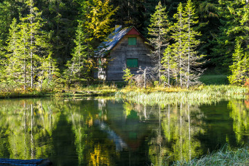 A small cottage at the side of alpine Joser Lake, located between the woods, next to a pathway leading to Buchbergkogel, Austria. Calm surface of the lake with many reflections. Forged perspective