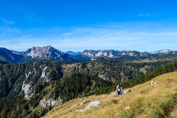 Fototapeta na wymiar A girl with a big hiking backpack walking through an Alpine meadow to the Buchbergkogel's top in Austria. There are endless mountain chains behind her. Struggle to achieve the goal. Early autumn vibes