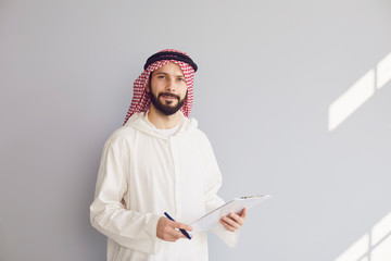 Attractive smiling arab man writes in clipboard on gray background