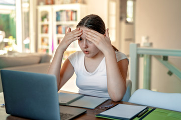 Teenager girl with shocked face studying online at home looking at laptop at quarantine isolation period during pandemic. Home schooling. Social distancing. Online school test. Overwhelming news.