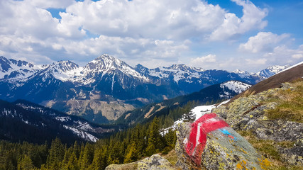 A path mark looking like flag of Austria painted on a big stone next to the pathway to Himmeleck peak, Austria. There is a massive mountain range in the back, partially covered with snow. Early spring