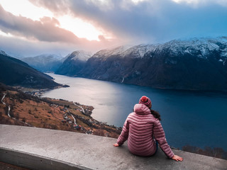 girl with brown hair and rose jacket sitting on the edge in noway watching the mountains