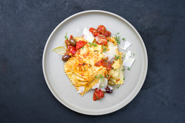 Traditional Italian ravioli pasta offered with parmesan cheese, fried tomatoes and olives as top view on a modern design plate with copy space