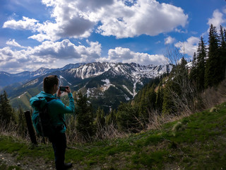 Naklejka na ściany i meble Woman with big hiking backpack taking a picture with a phone of the landscape, while hiking to Himmeleck peak, Austrian Alps. There is a massive mountain range in the back, partially covered with snow