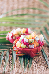 Exotic fruit salad served in half of dragon fruit over palm leaves on rattan background. Copy space. Tropical travel, exotic fruit. Vegan and vegetarian concept.
