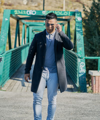 portrait of a handsome Hispanic boy walking on a metal pedestrian bridge with good pants and urban clothing: jeans, blue shirt and dark jacket and sunglasses on the face