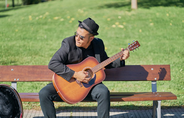 Hispanic male musician playing acoustic guitar sitting on a park bench with his black hat and sun glasses